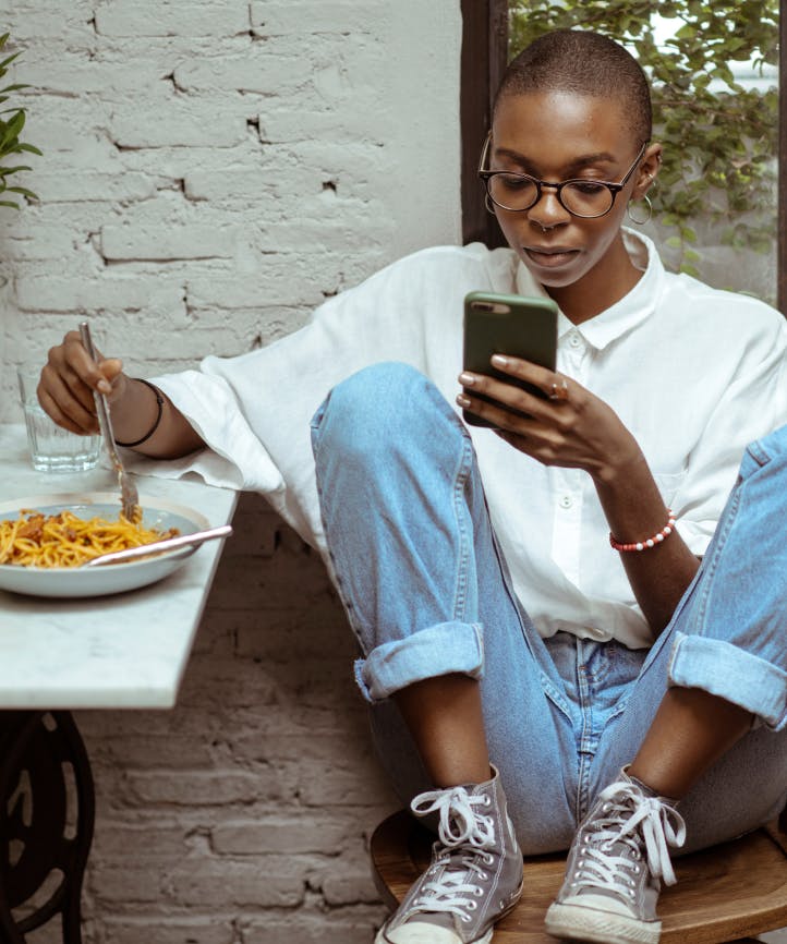 A woman checks her phone while casually eating a pasta meal.